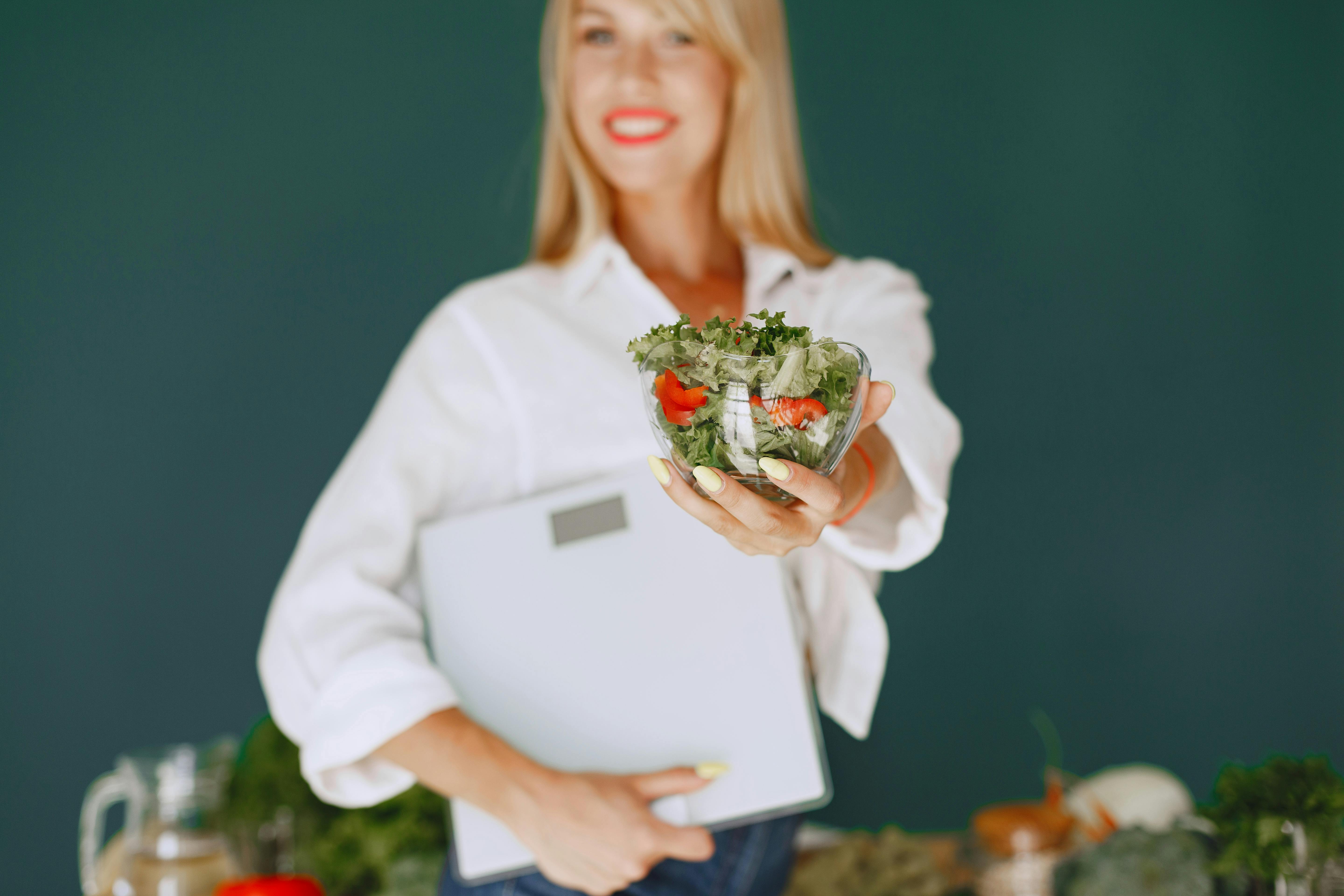 A woman, standing in front of a chalkboard, holding a clipboard, and showing you a small bowl of vegetables.