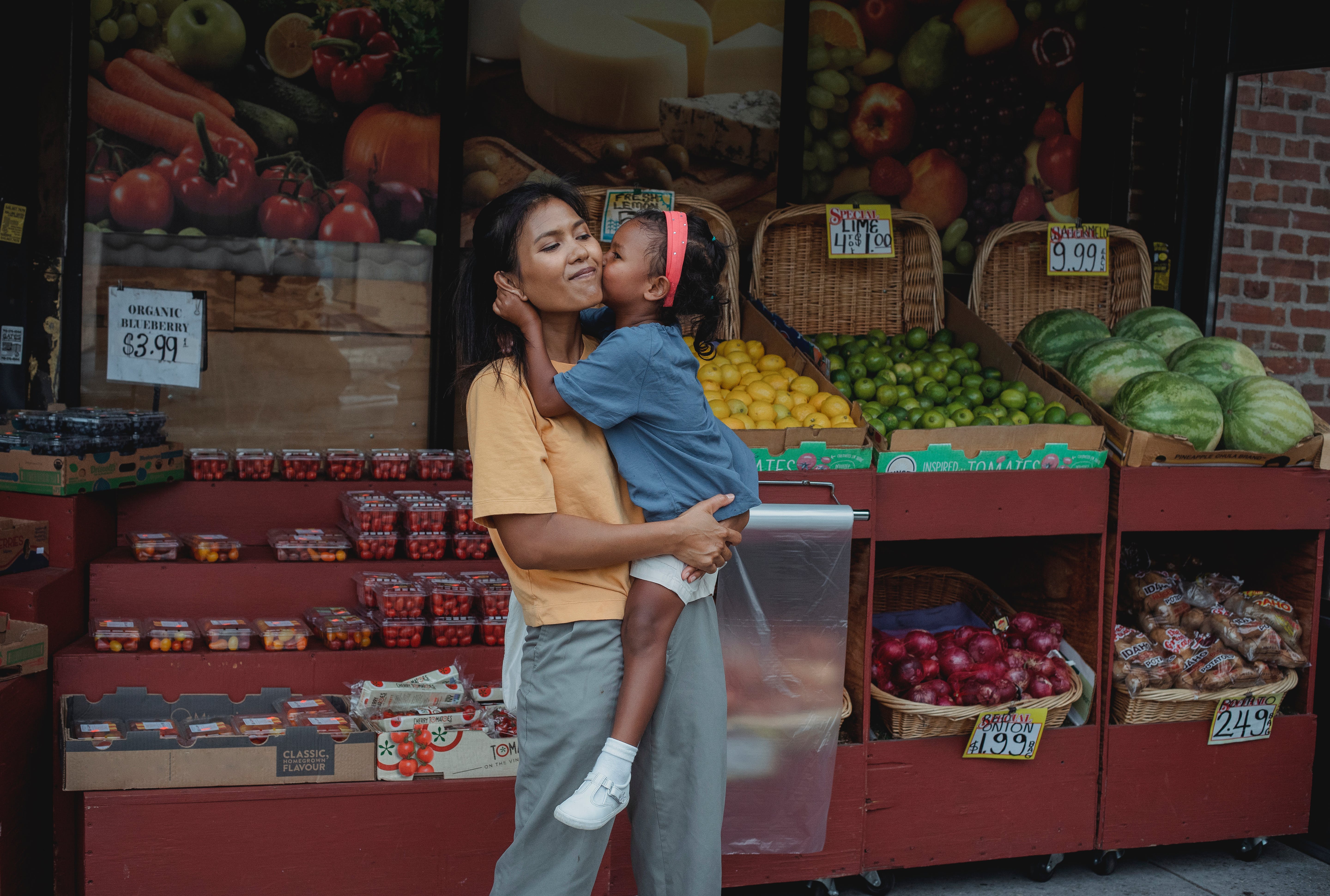 A woman picking up, and being kissed by her child, in front of the vegetables being sold.